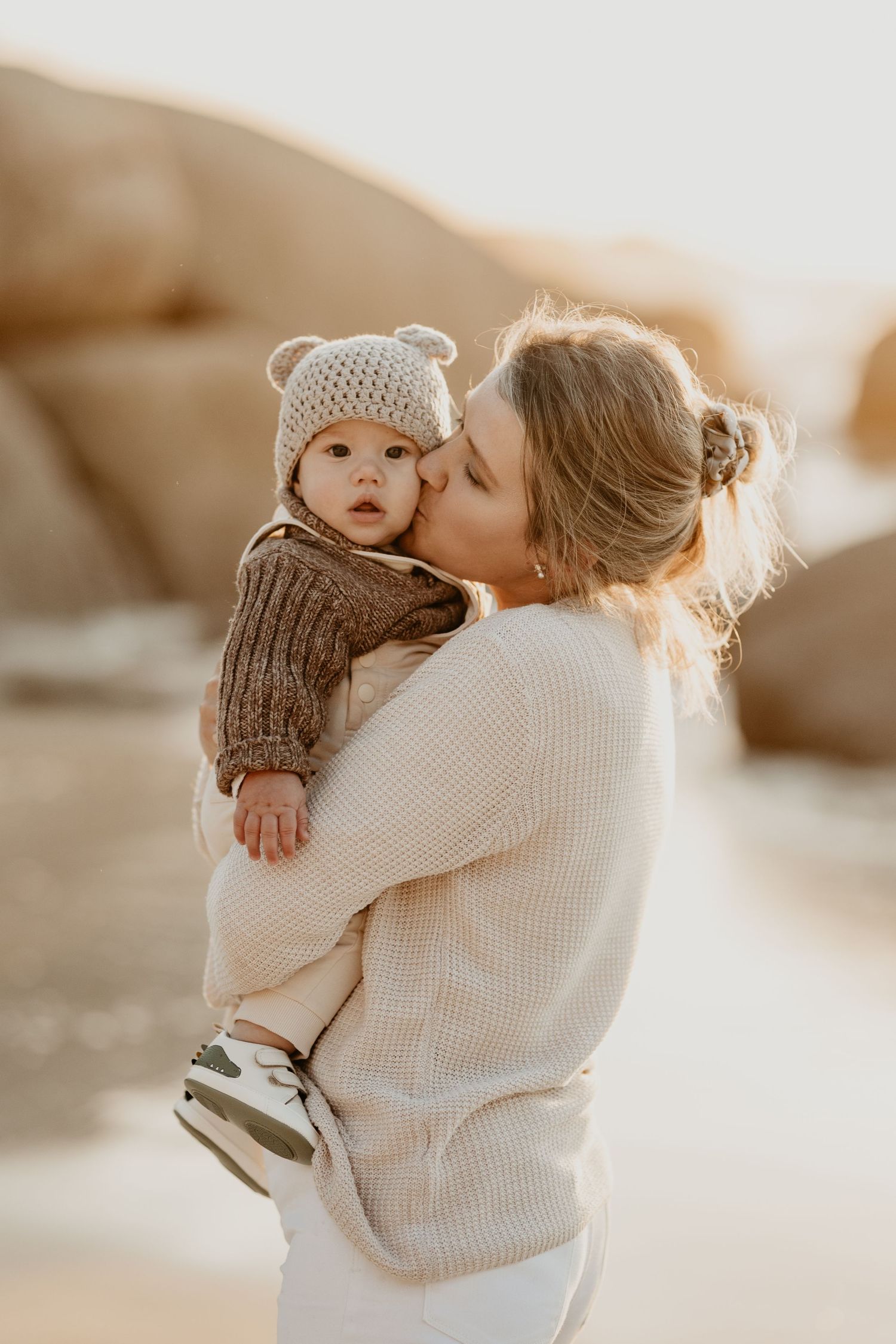 Parent in cream sweater holds and kisses baby wearing knit hat and mittens during golden hour outdoor photoshoot.