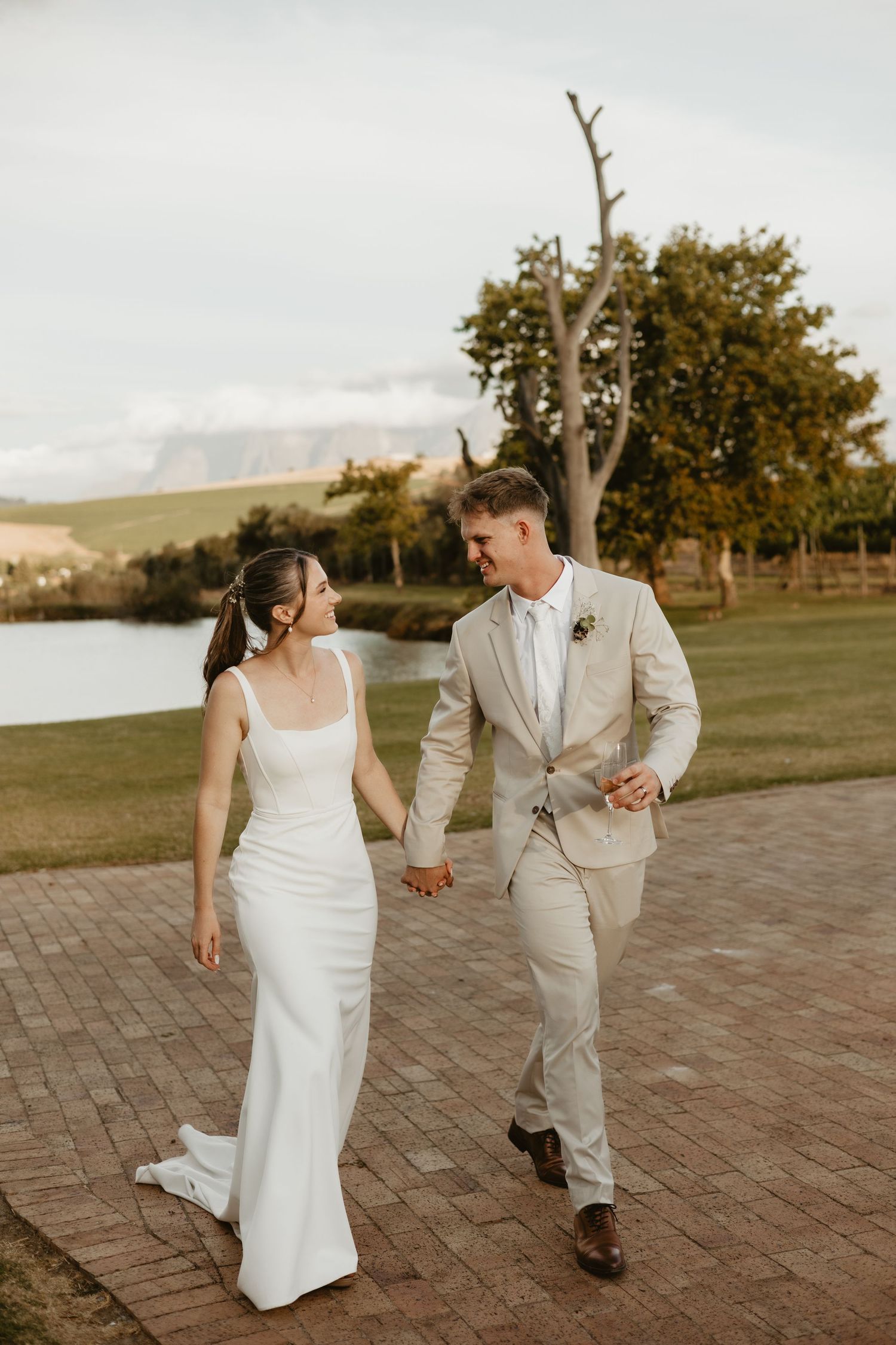 A couple in wedding attire walks hand in hand on a brick path by a lake during golden hour at sunset.