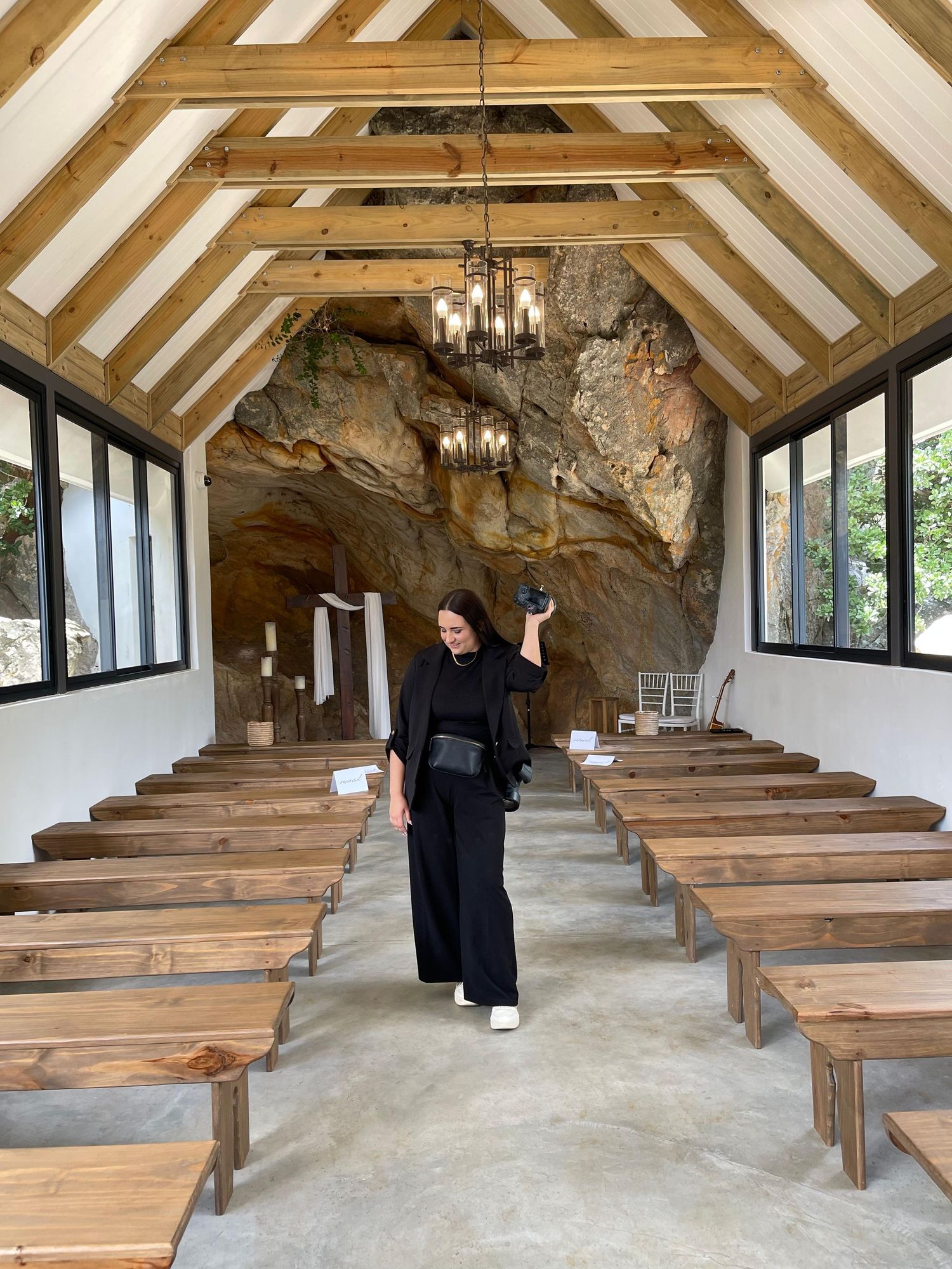 Woman in black outfit holding camera in rustic cave chapel with wooden pews and chandelier.