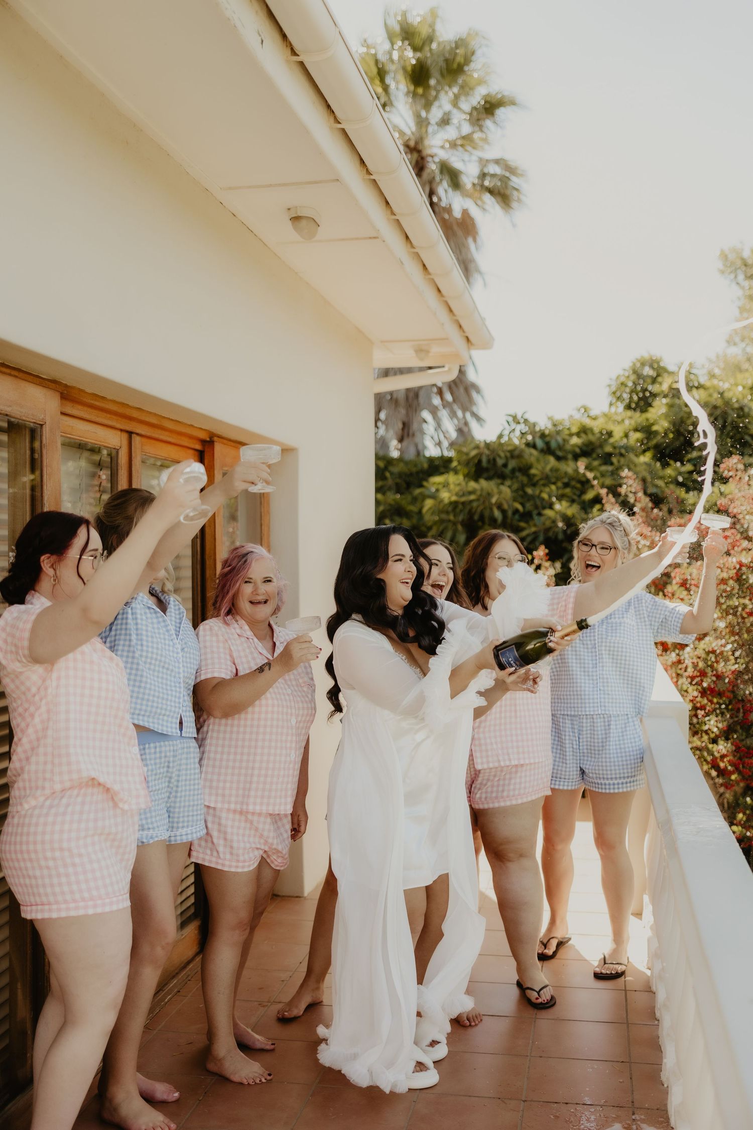 Bride in white gown pops champagne with bridesmaids in pink gingham pajamas on a sunny villa balcony.