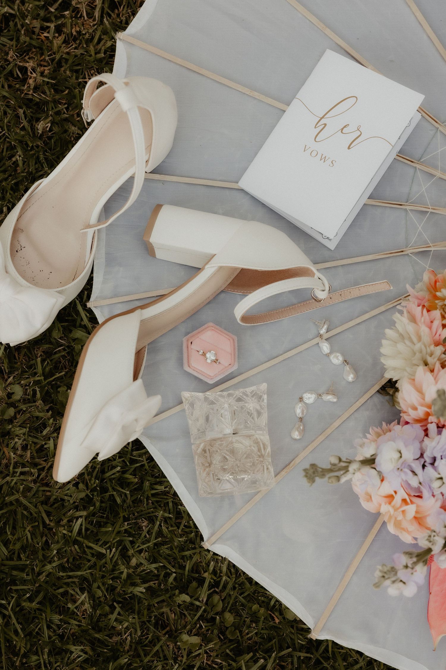 Bridal flat lay with white heels, vow book, pink ring box, lace garter, pearl earrings, and floral bouquet on white cloth.