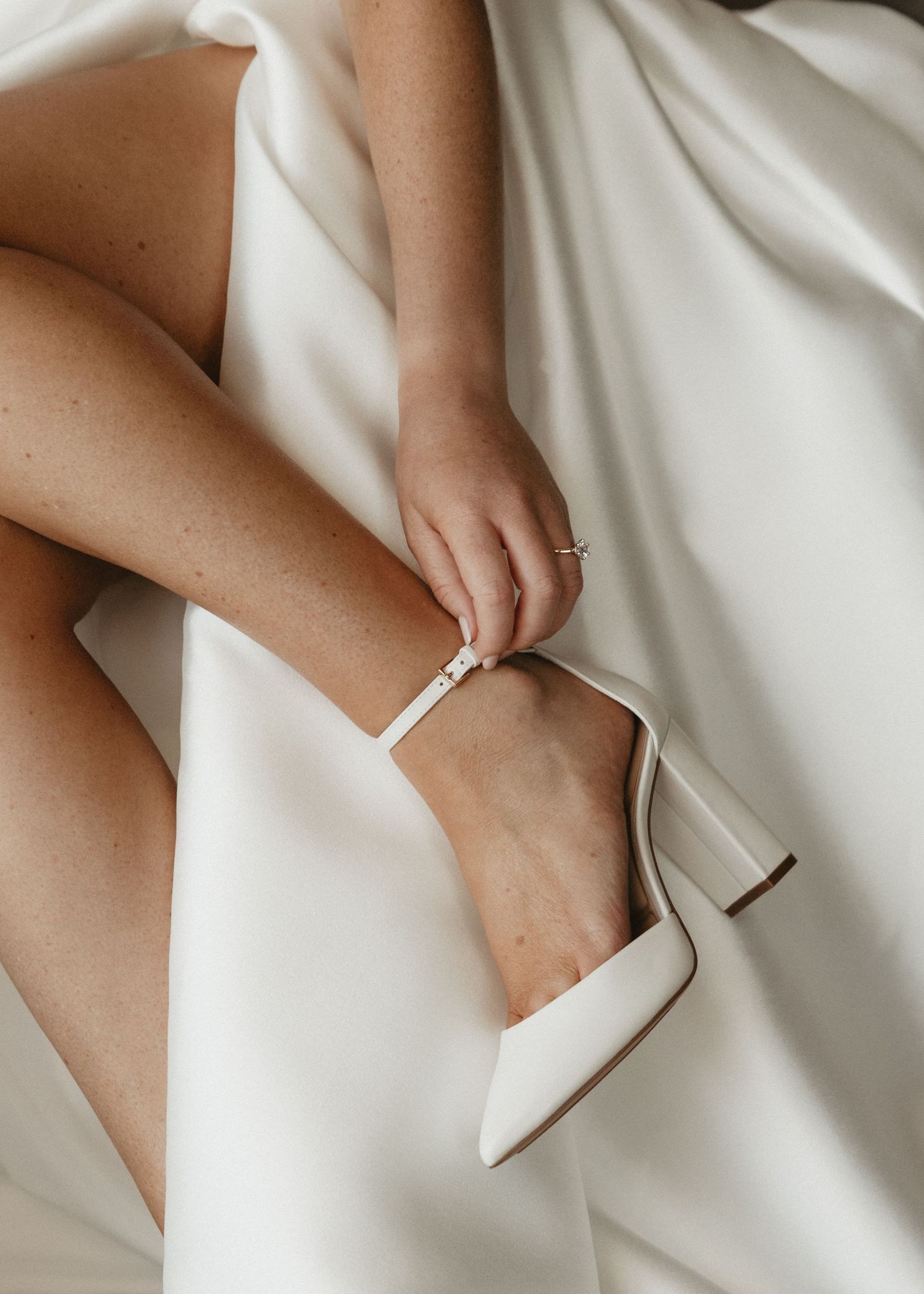 Close-up of a white bridal heel being fastened on a foot against white fabric.
