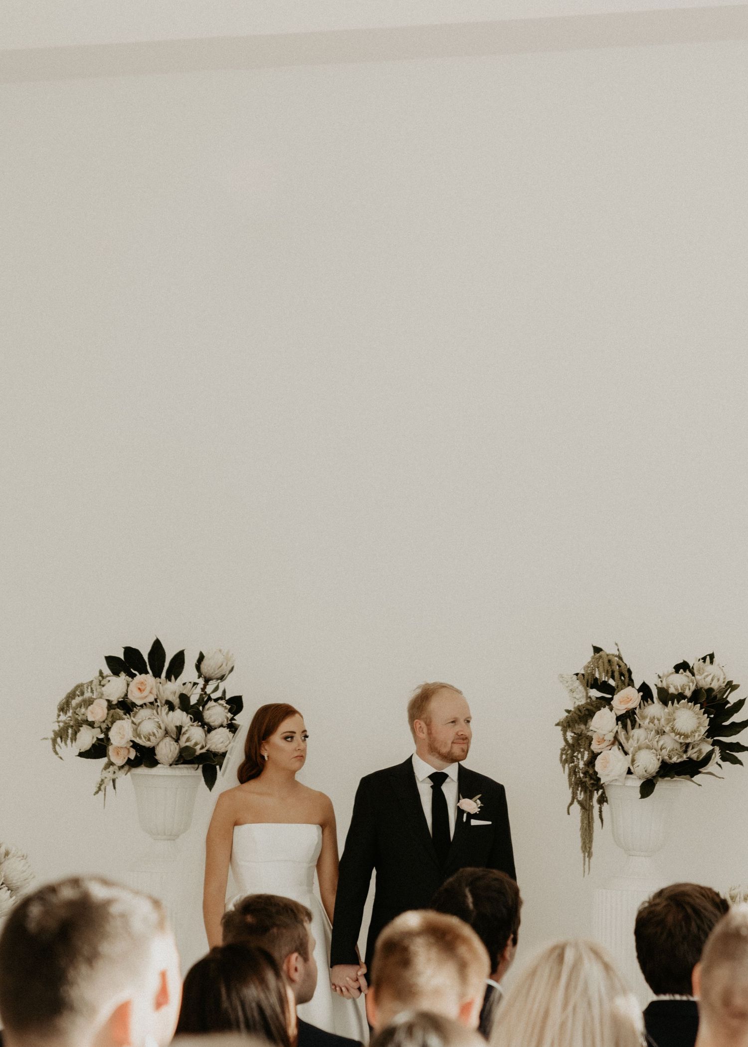 A bride and groom stand before their wedding guests with elegant white floral arrangements.