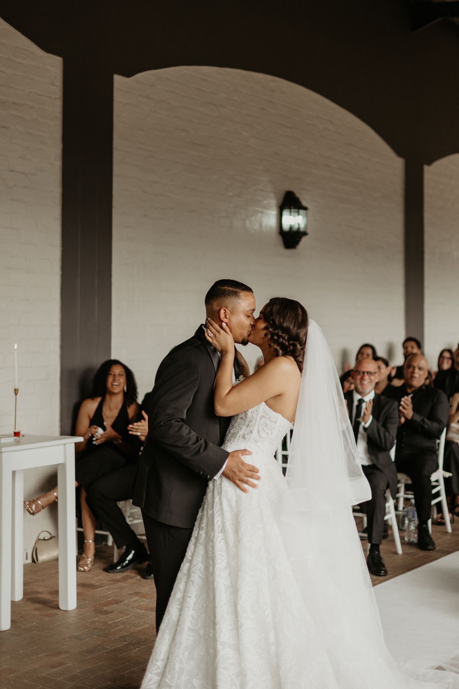 A romantic wedding couple shares a tender kiss while guests celebrate in a minimalist white venue with pillars.