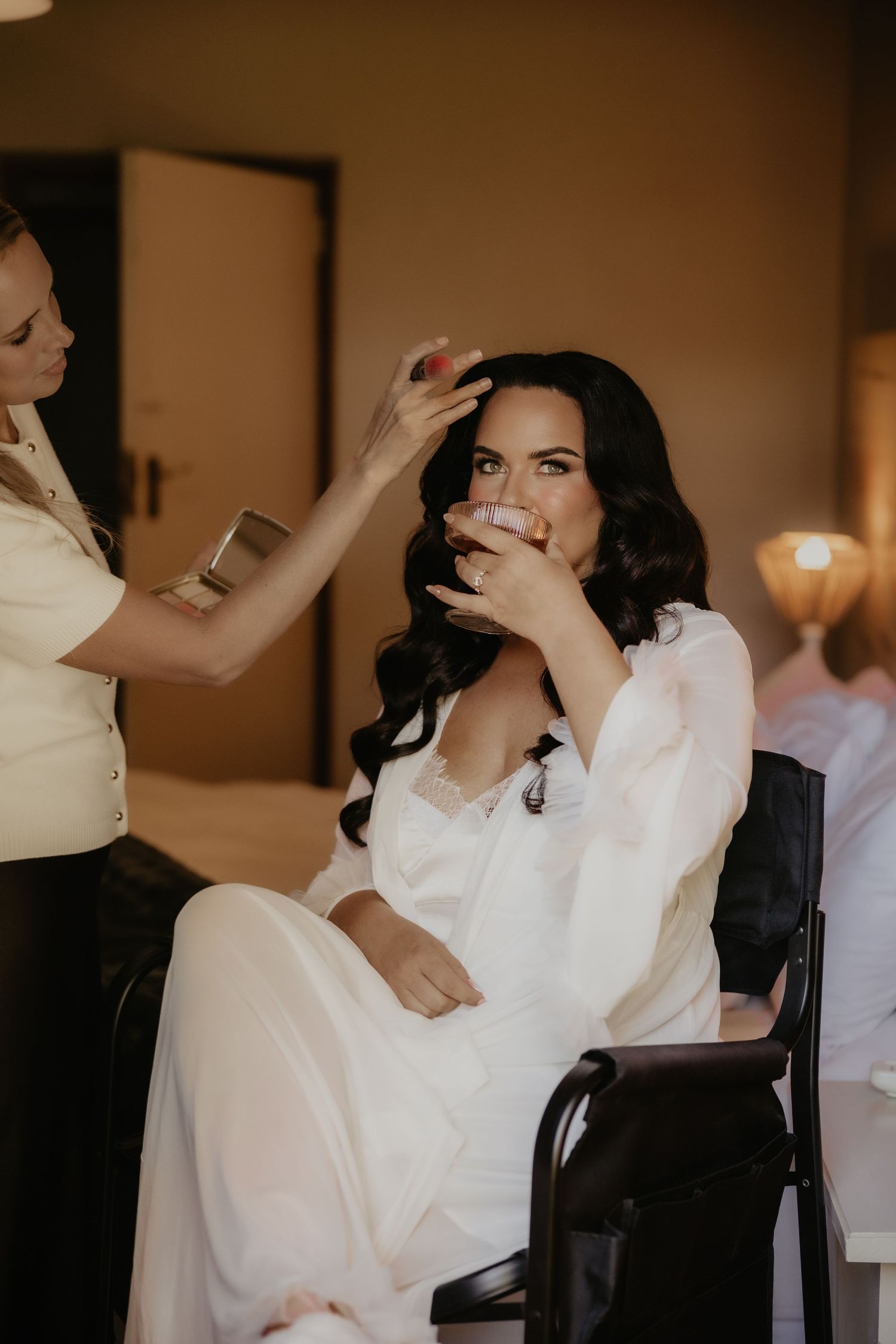 Bride in white robe sipping champagne while makeup artist applies finishing touches during wedding morning preparations.