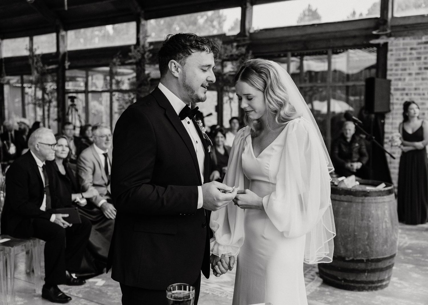 Wedding reception guests mingle around wine barrels in rustic indoor venue.