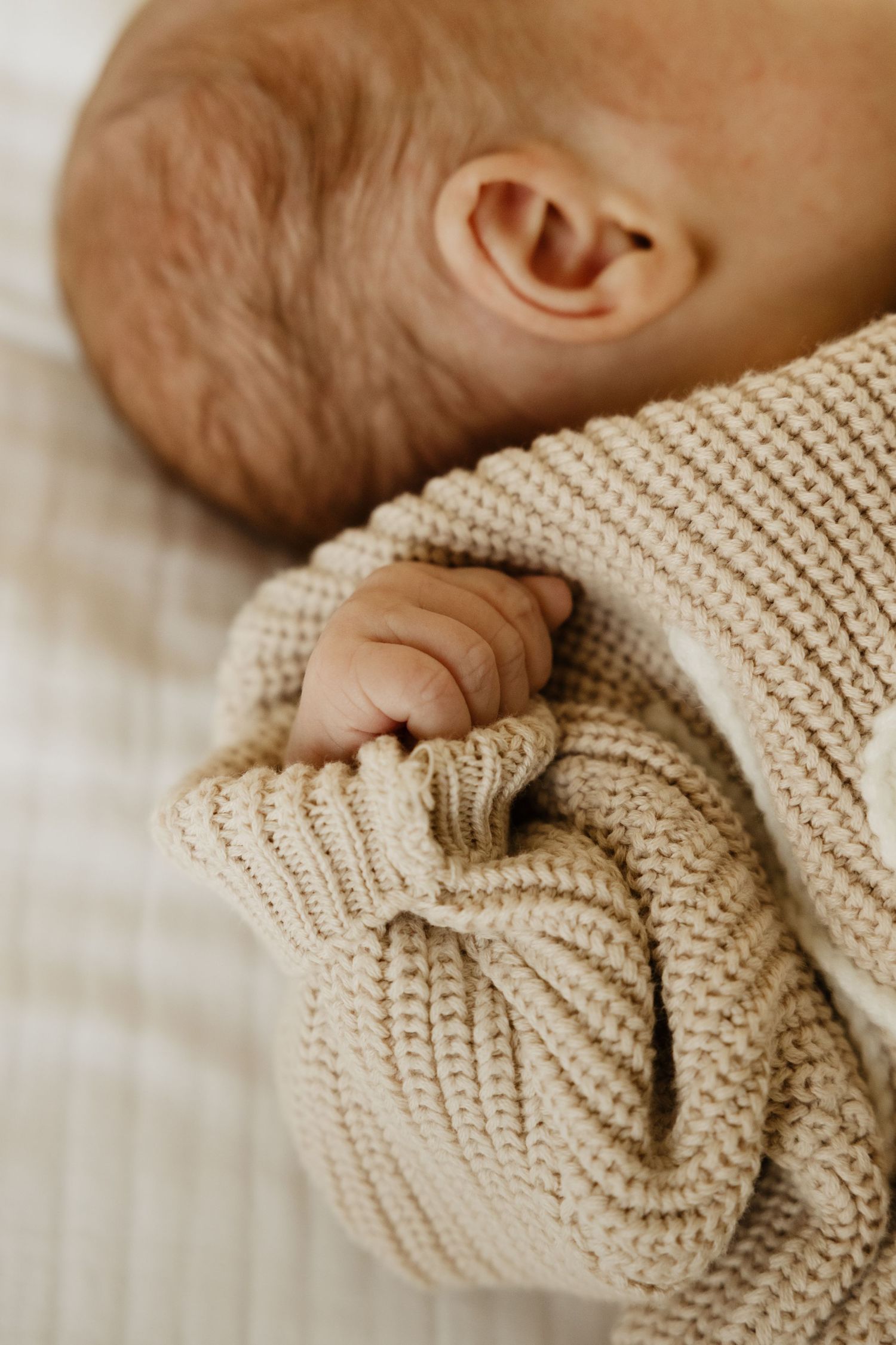 Close-up of a newborn baby in a beige knit sweater, tiny hand gripping the fabric while lying on a soft blanket.