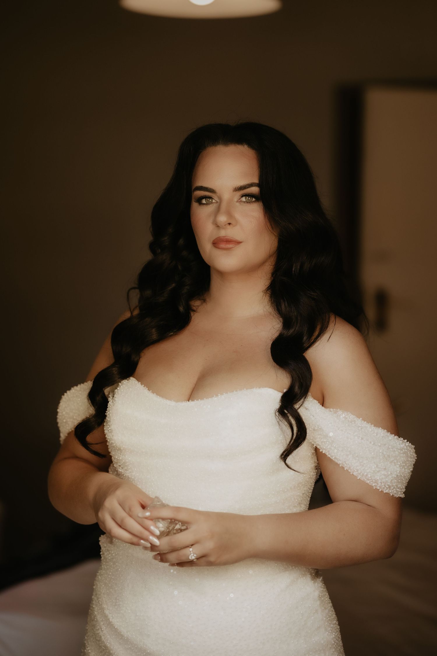Bride in white off-shoulder beaded gown with dark curly hair holding a glass, posing elegantly indoors.