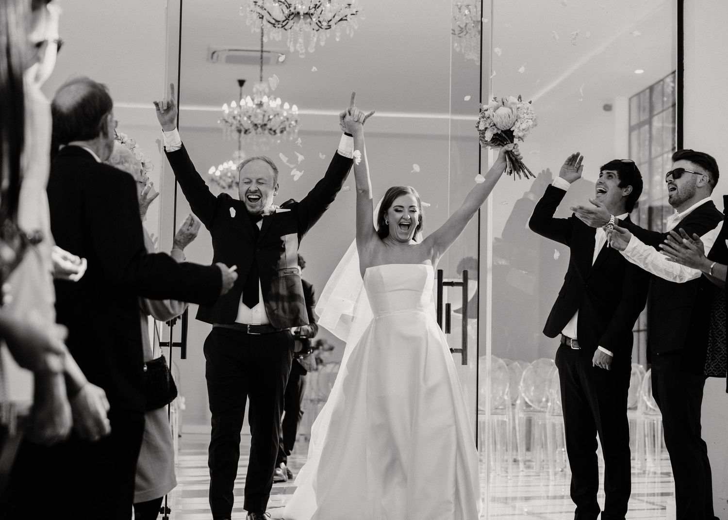 Wedding party celebrates joyfully under a chandelier during a black and white reception moment.