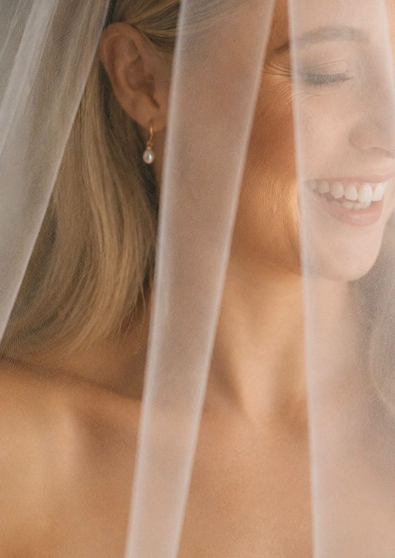 Smiling bride wearing pearl drop earrings, partially covered by a sheer white wedding veil in a soft, romantic portrait.