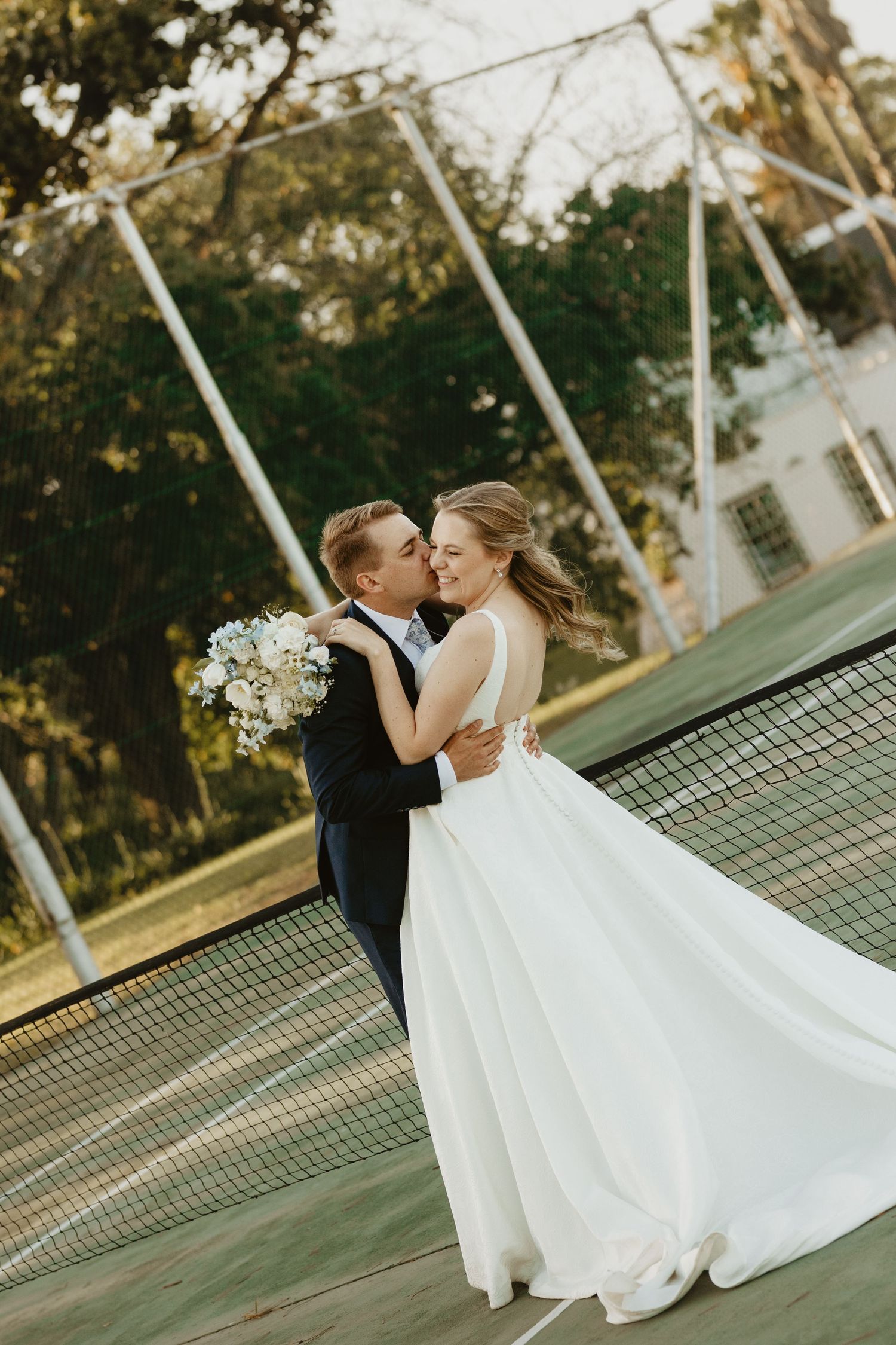 A romantic wedding couple shares a tender moment on a tennis court at sunset.
