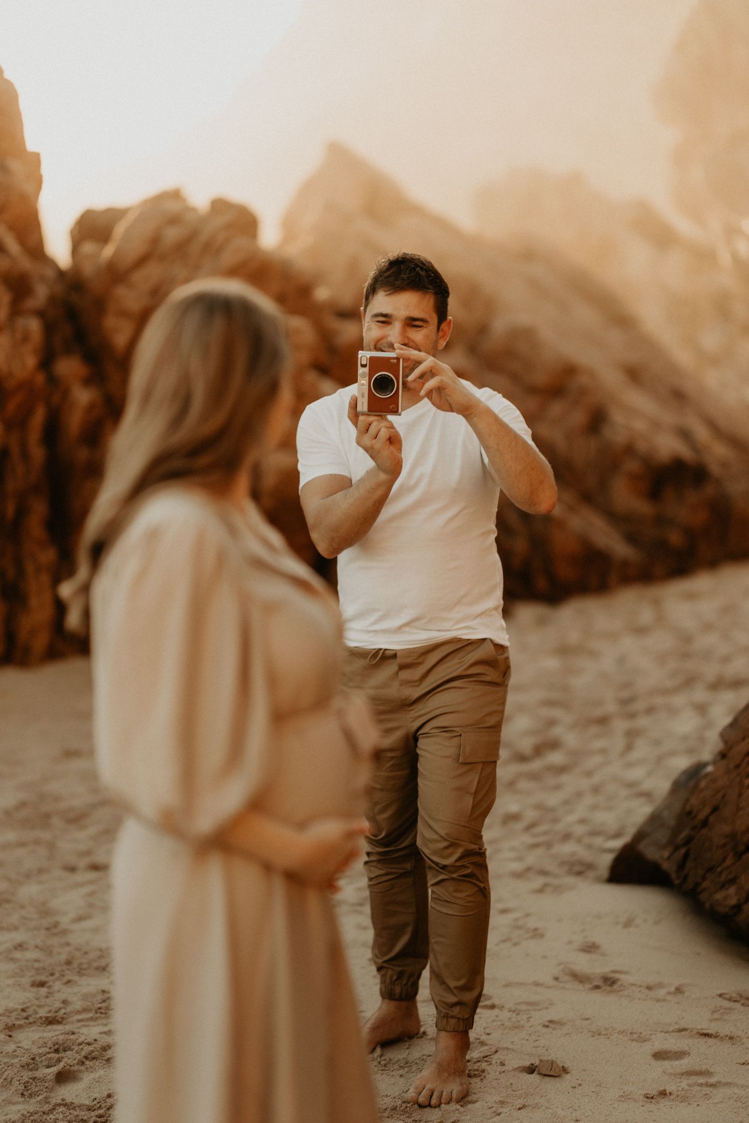 A couple shares a romantic moment on a sandy beach with dramatic rock formations at sunset.
