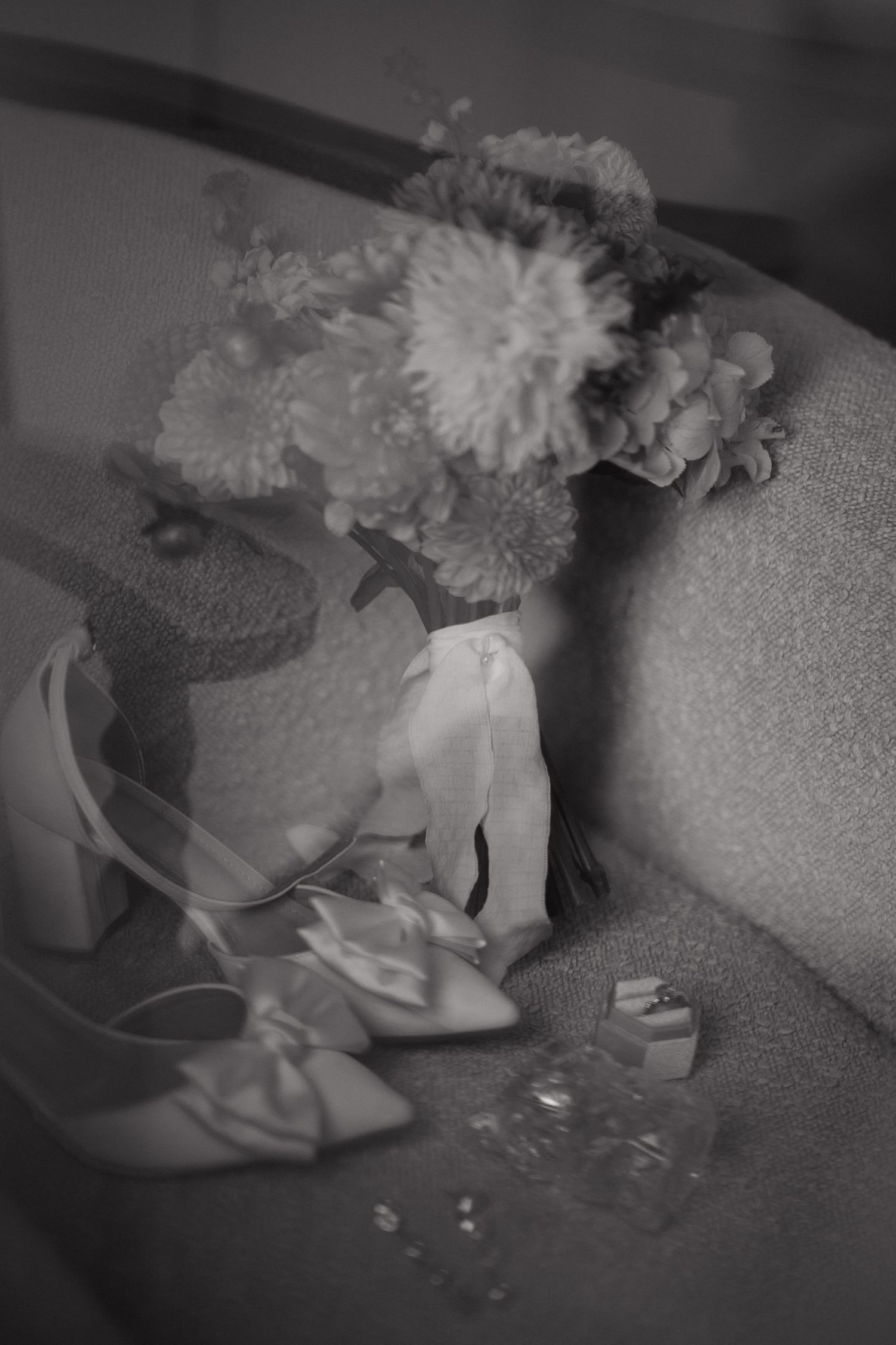 Black and white photo of bridal accessories including heels, bouquet, and jewelry arranged on a sofa.