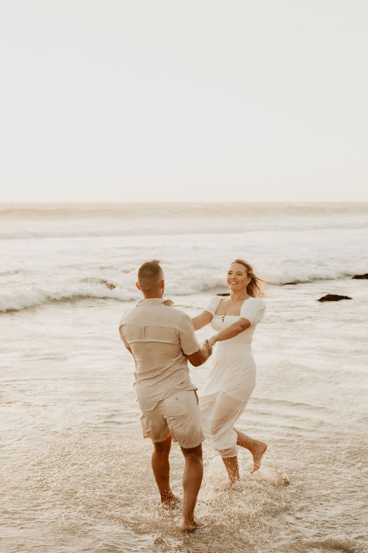 A couple in light colored clothing plays joyfully in the ocean waves at sunset, creating a romantic beach scene.