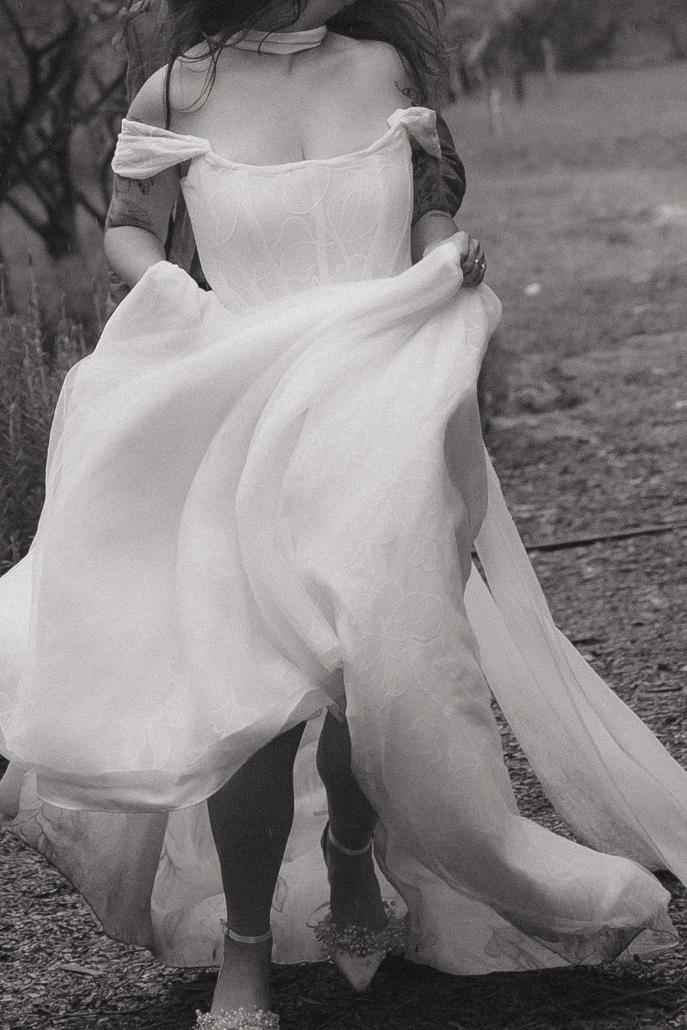 Black and white photo of a bride in an off-shoulder wedding gown, skirt flowing in the wind outdoors.
