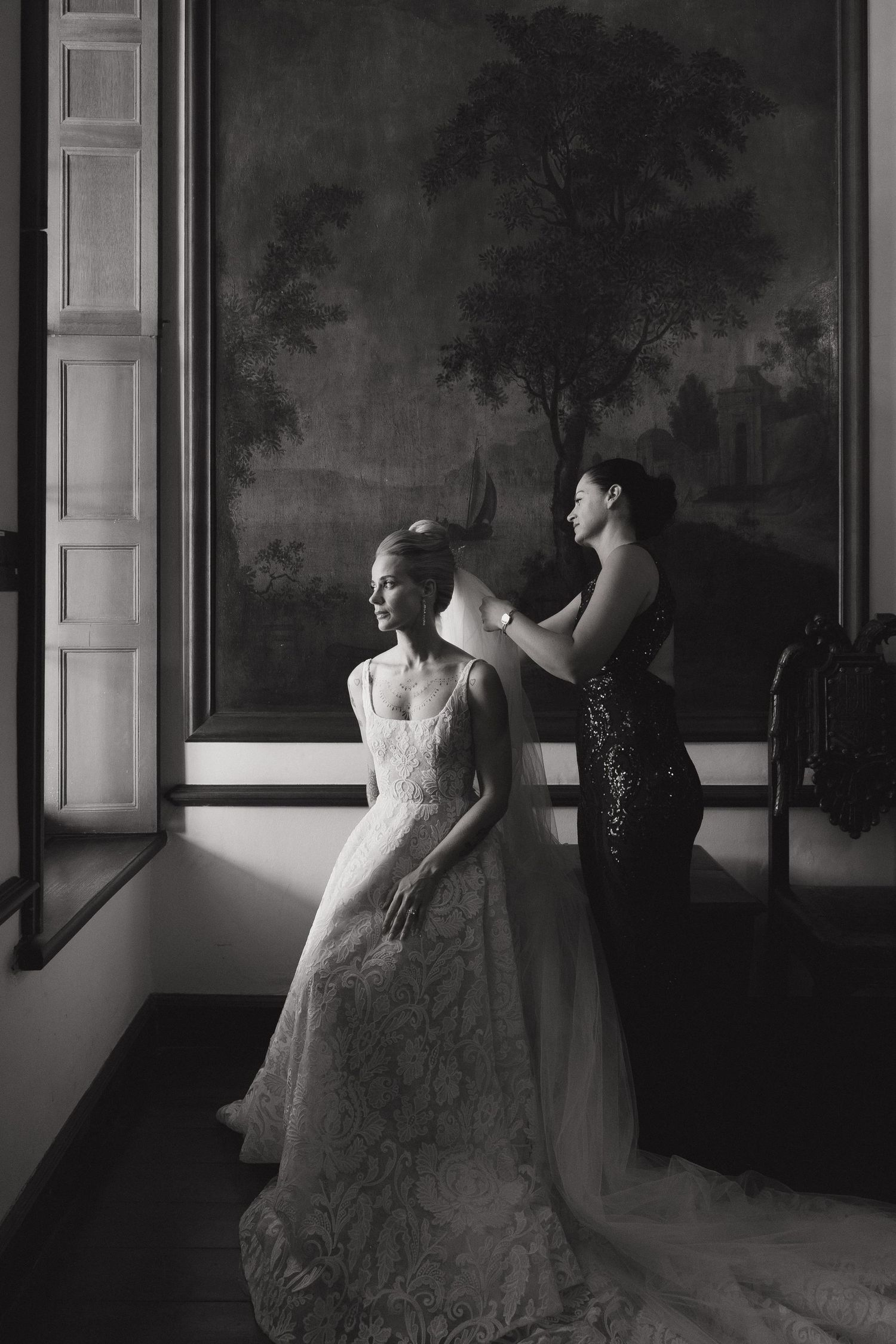 Black and white photo of bride in lace gown being assisted by bridesmaid before wedding ceremony.