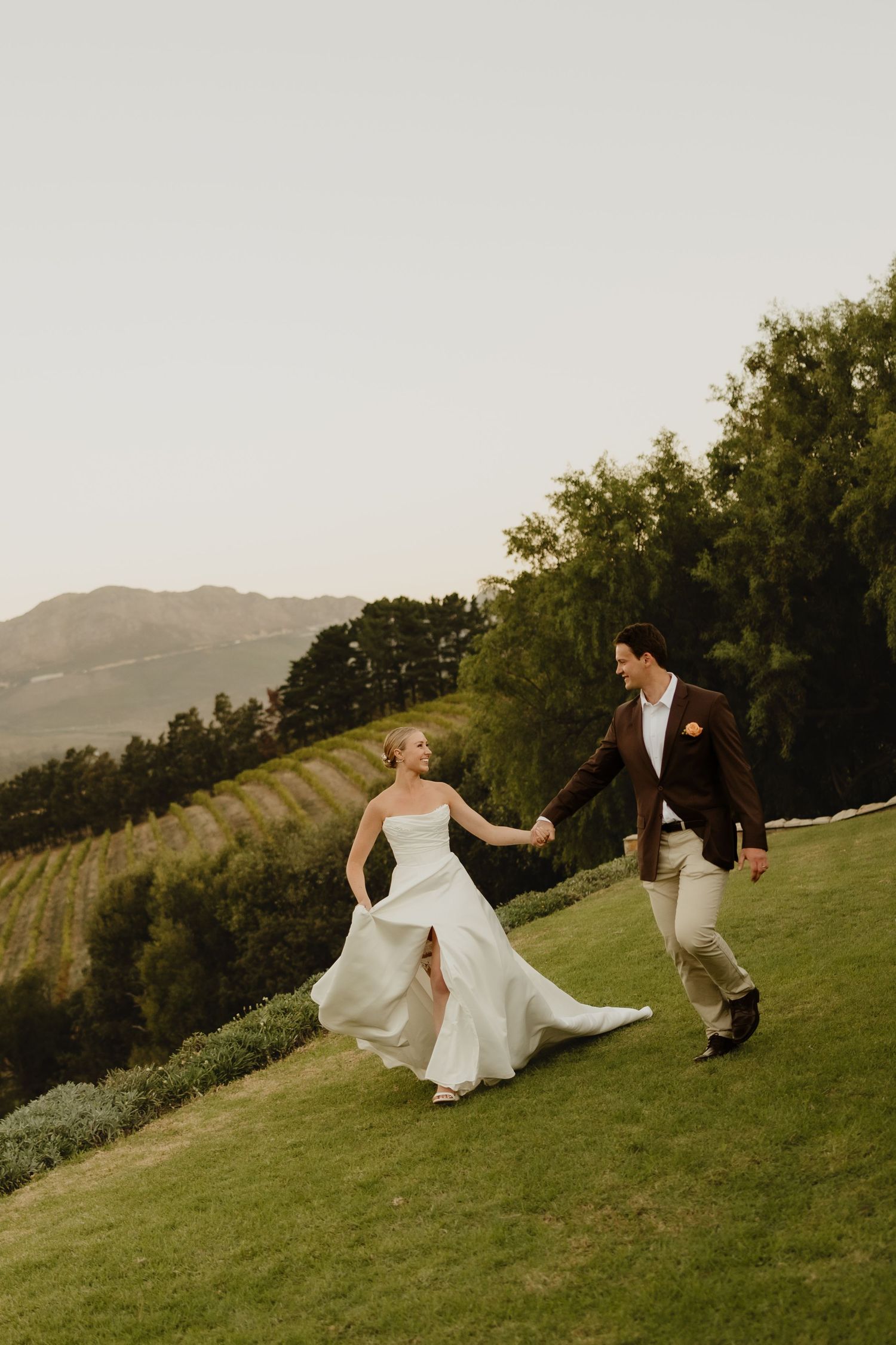 A bride in a white gown and groom in a brown suit hold hands while walking on a green hillside with vineyard views.