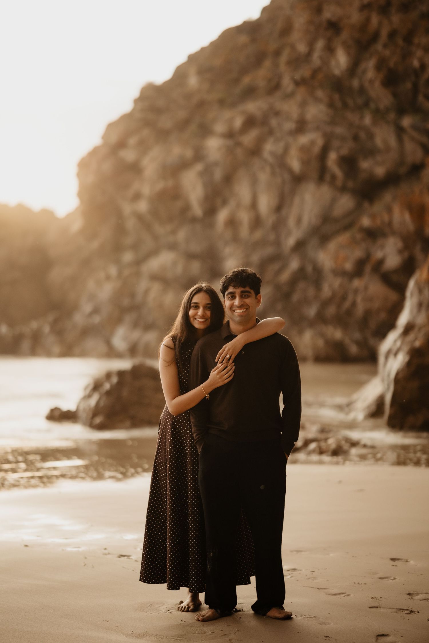 Couple embracing on a sandy beach at golden hour with dramatic rocky cliffs in the background.