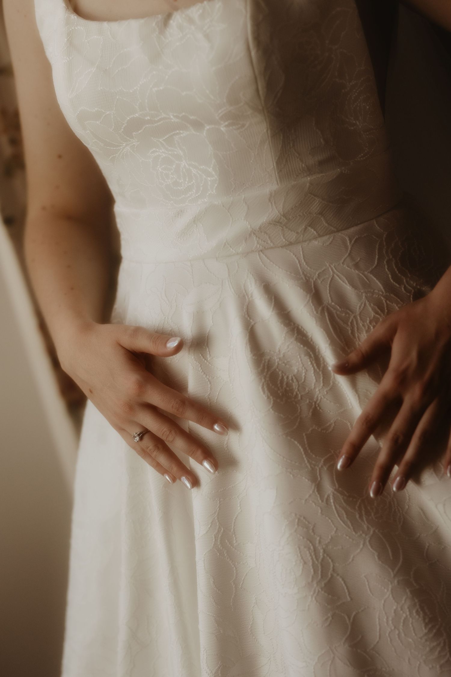 Close-up of a bride in a white floral lace wedding dress, hands resting on waist, wearing an engagement ring.