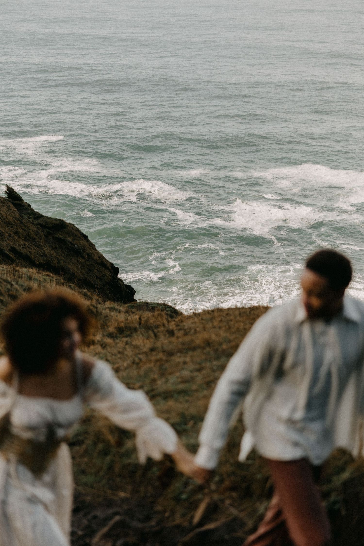 Couple holding hands while walking along dramatic coastal cliffs with crashing waves below.