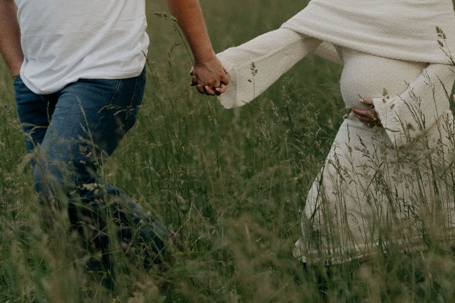 Romantic couple walking through tall grass field in white flowing clothing.