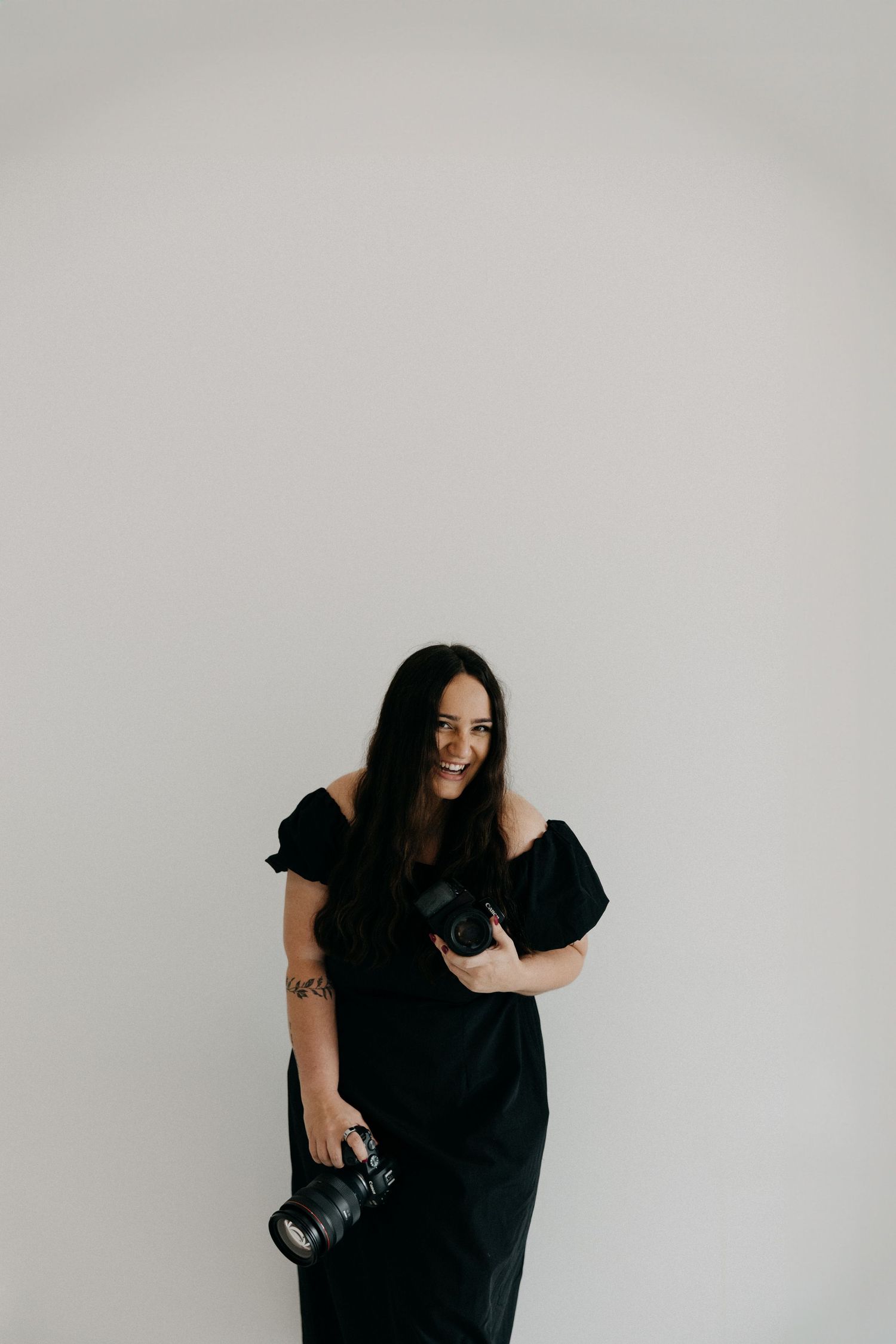 Professional photographer in a black dress holds a camera while posing against a white studio backdrop.