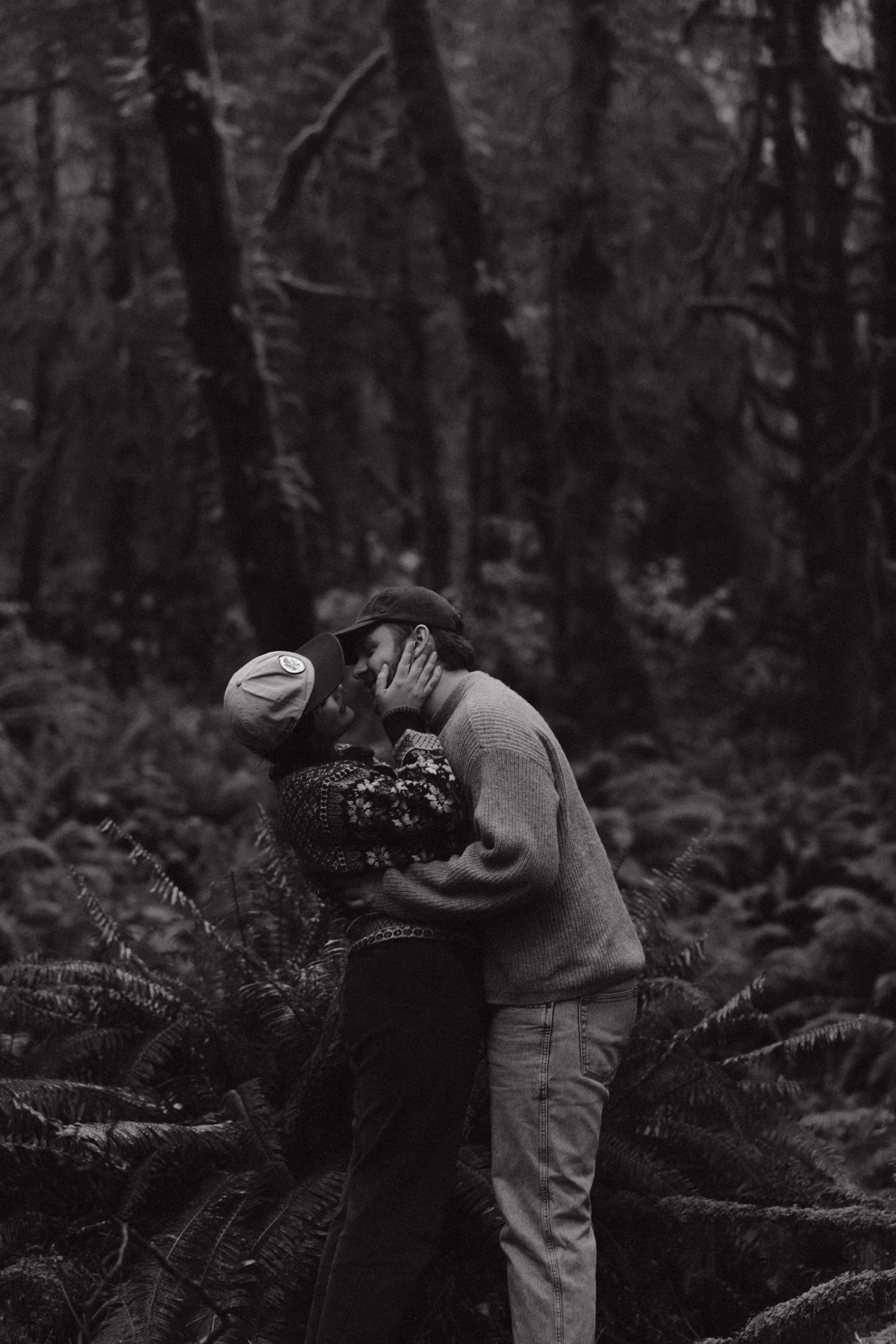 A romantic black and white photo of a couple embracing in a moody forest setting among ferns and trees.