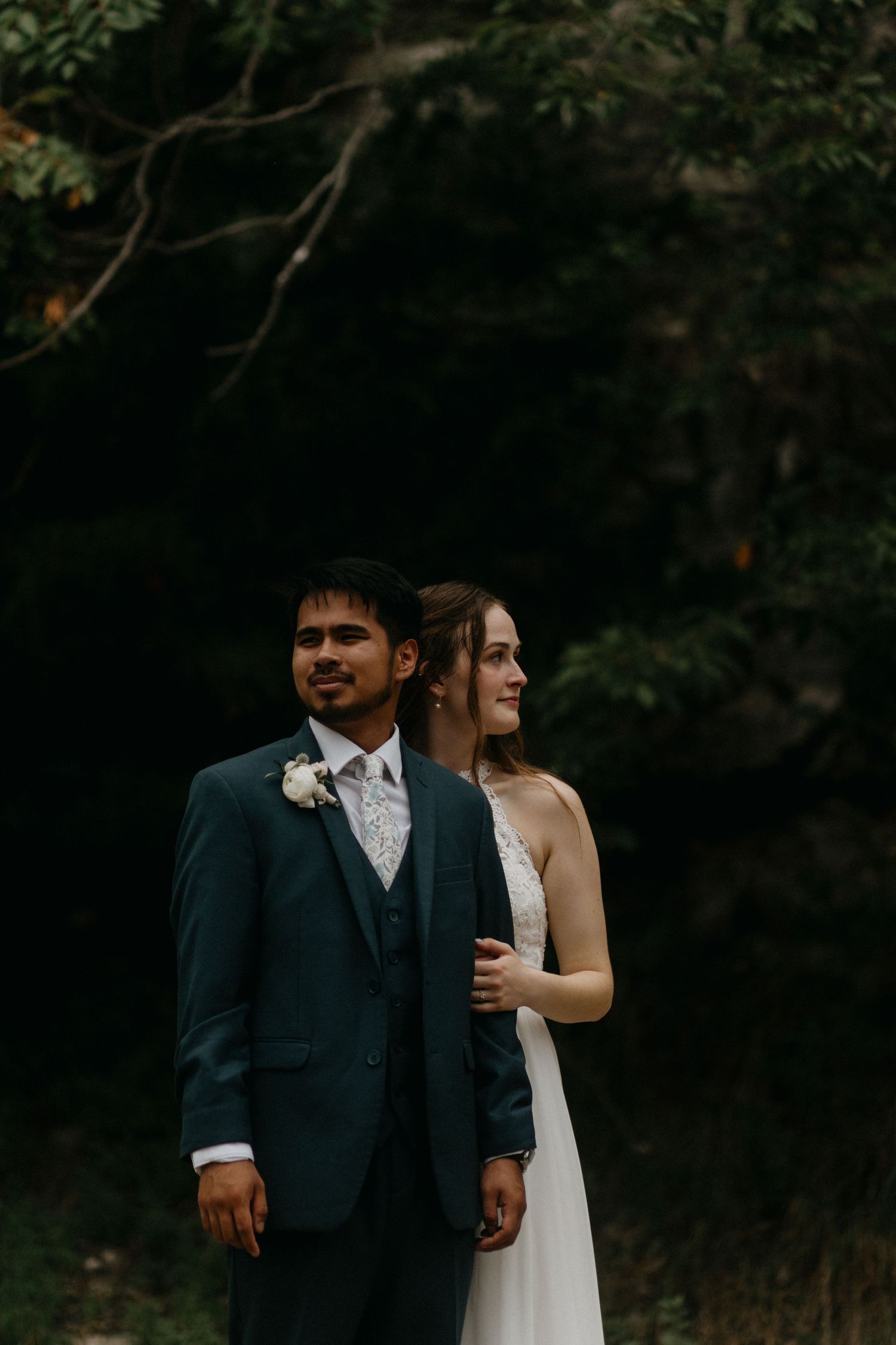 A couple in wedding attire stands together in a moody outdoor forest setting at dusk.