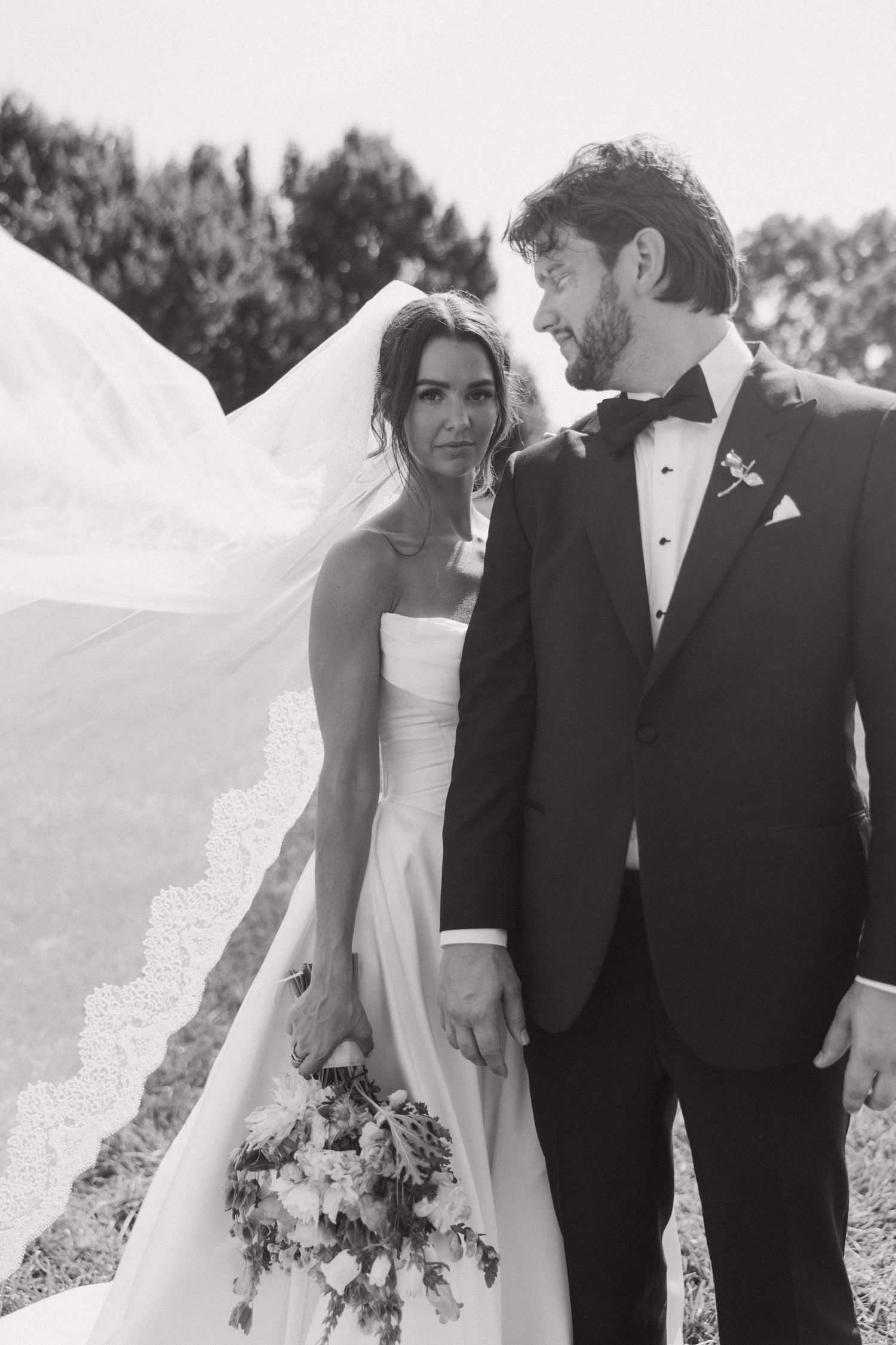 A romantic black and white wedding photo capturing a sweet moment between newlyweds with a flowing veil and floral bouquet.