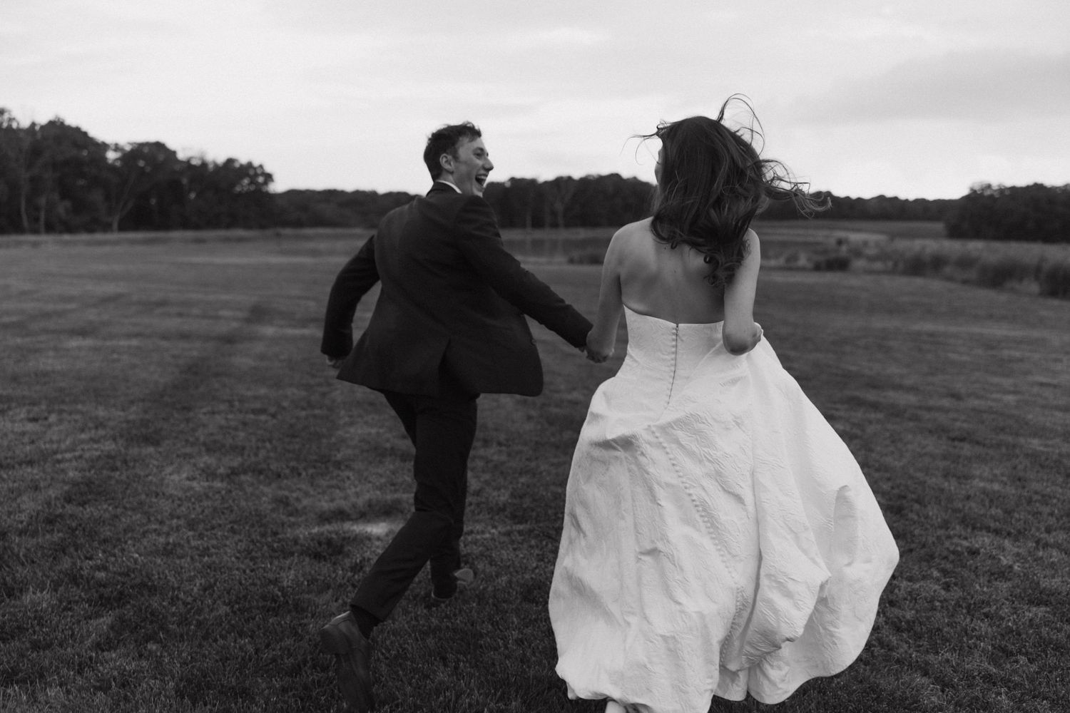 A couple in wedding attire runs playfully across an open field on their special day, captured in black and white.
