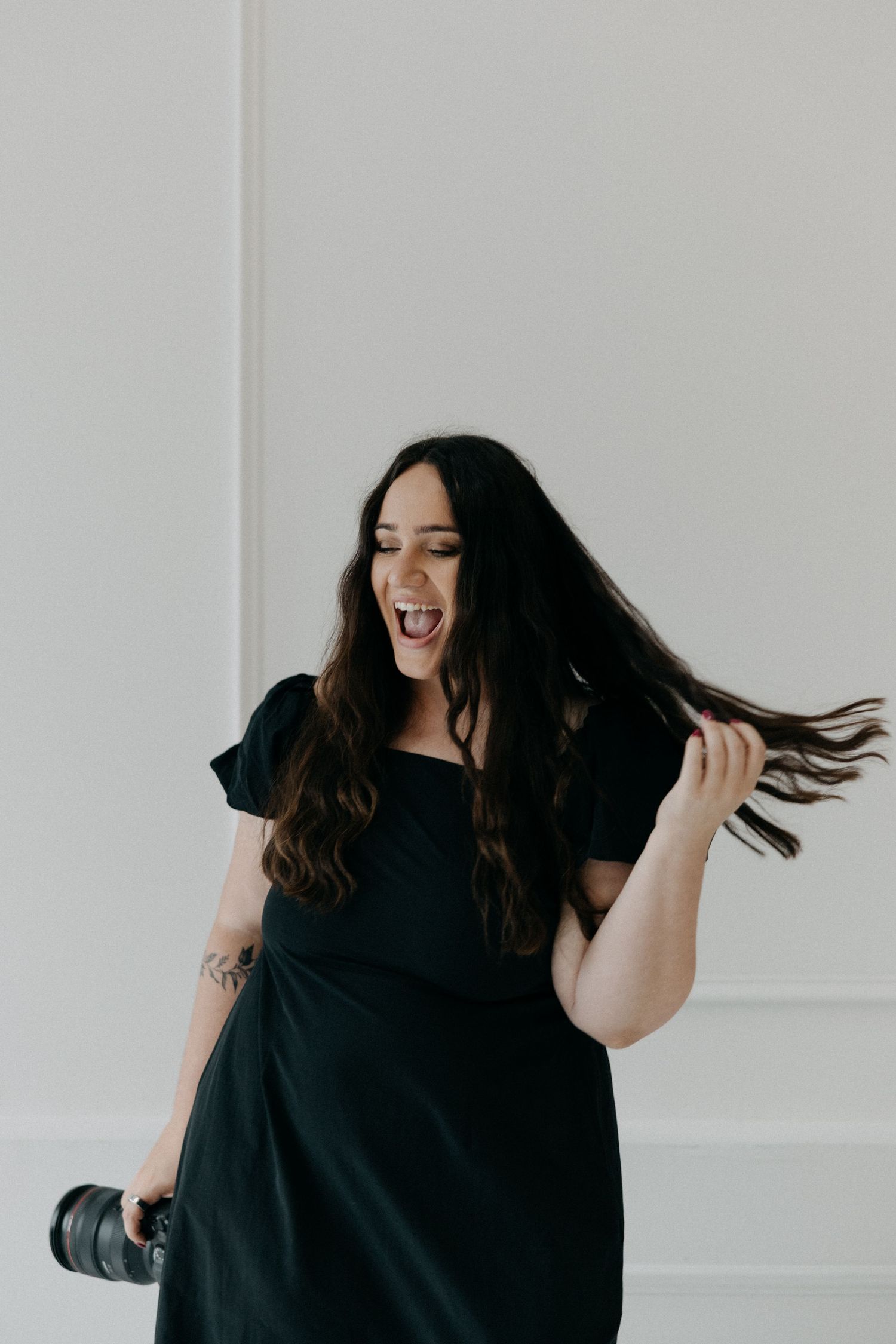 An energetic woman in a black dress dances against a white wall while holding a camera lens.