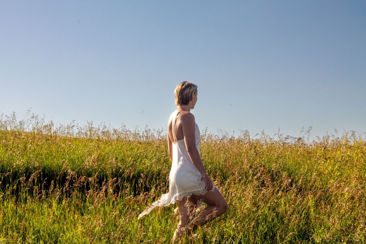 A woman in a white dress walks through a sunny green meadow with tall grass under a clear blue sky.