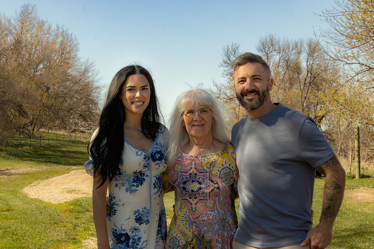 Three people smiling outdoors in a sunny park with trees, an elderly woman flanked by a young woman and a man.