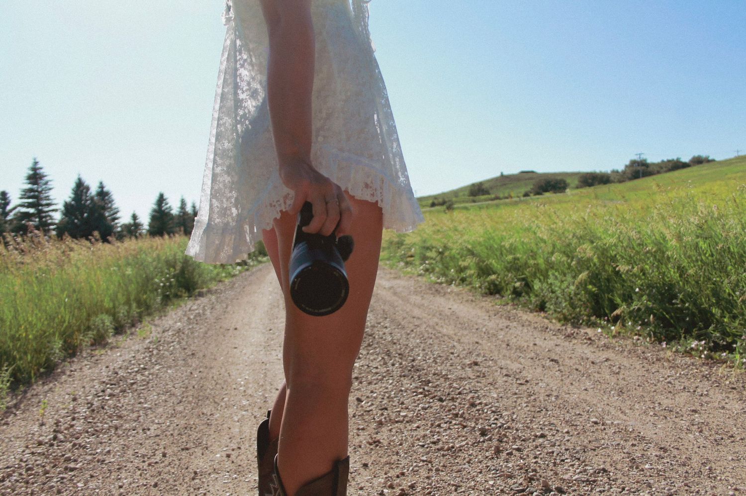 Person in white dress walking on dirt path through sunny summer landscape.