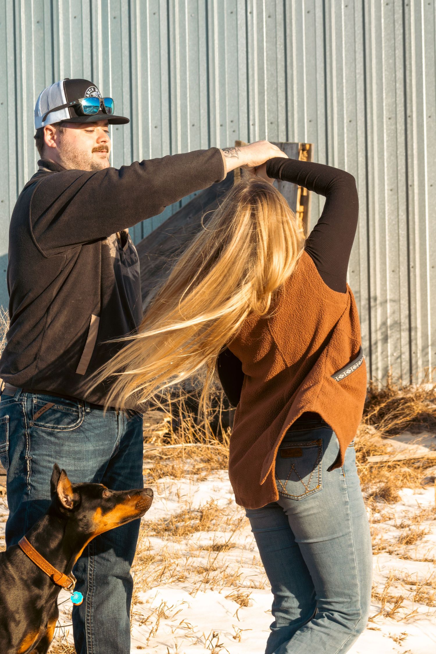 Two people in casual attire playfully interact outdoors in winter with a dog wearing an orange collar nearby.