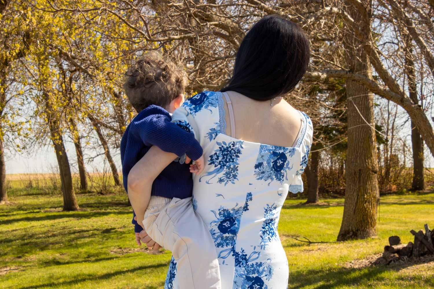 Woman in floral dress holding a young child outdoors in a sunny park with autumn trees.