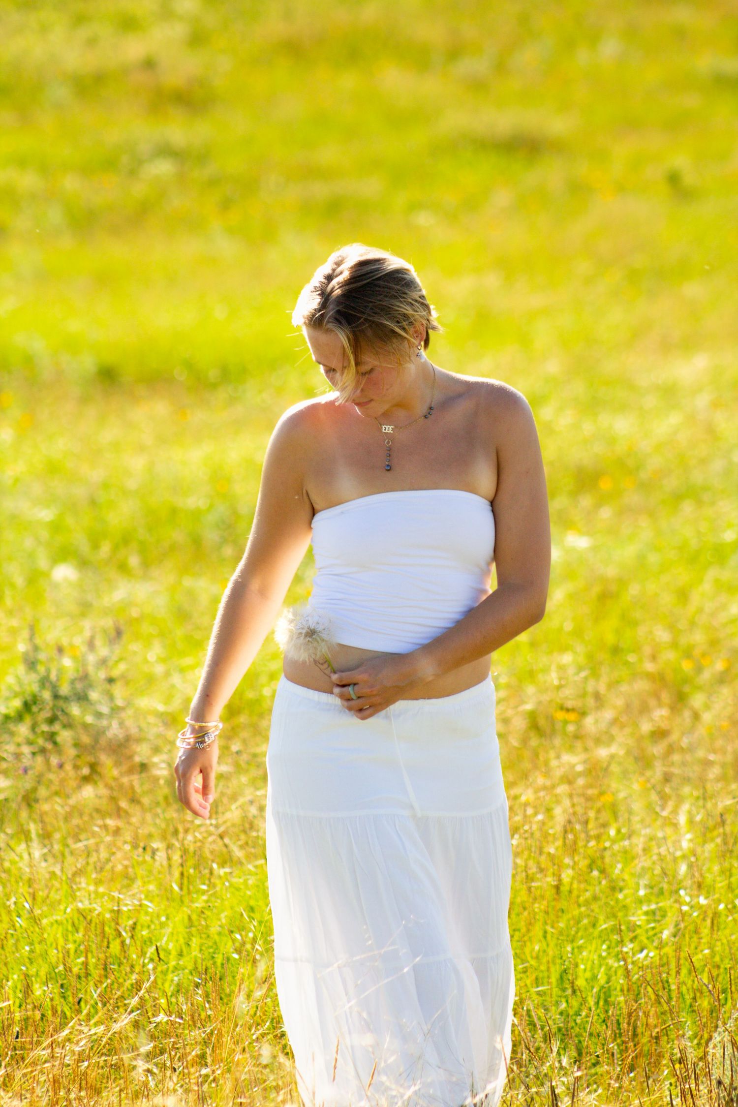 Pregnant woman in white strapless dress standing in sunny green field, cradling her baby bump with a peaceful downward gaze.