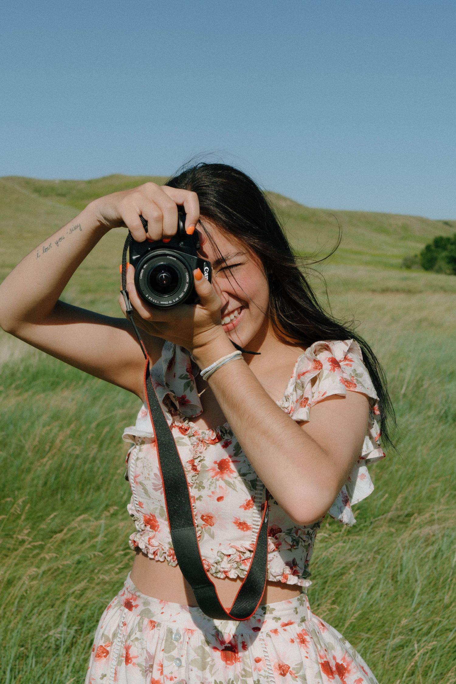 Person in floral dress taking photos with a camera in a grassy meadow on a sunny day.
