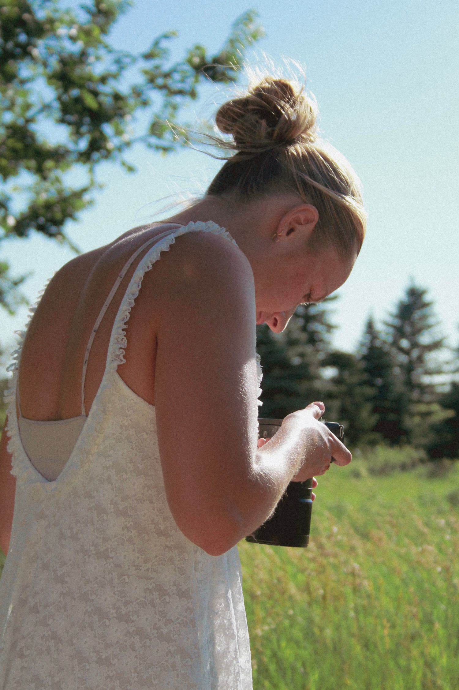 A photographer in a white lace dress examining their camera on a sunny summer day in the countryside.