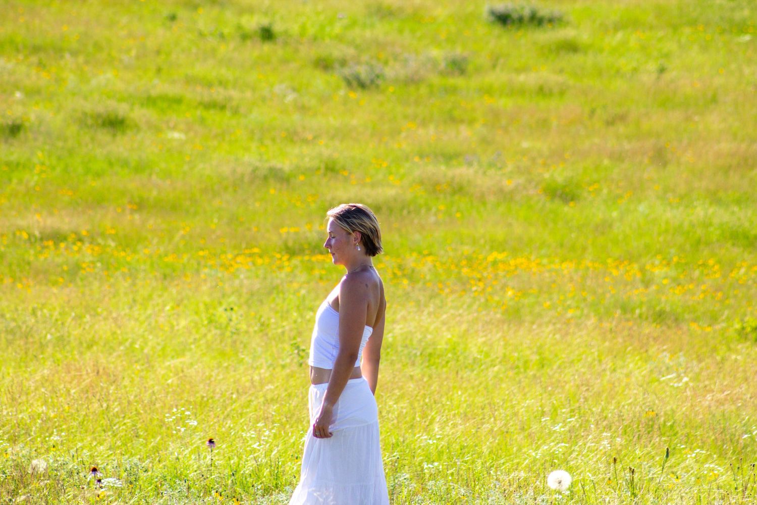 A woman in a white dress walks through a sunny green meadow filled with wildflowers.