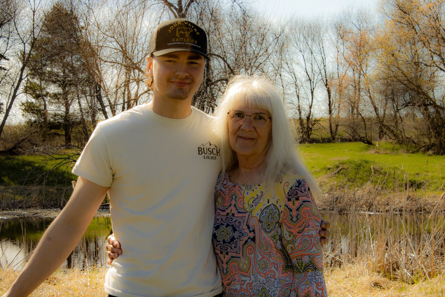 A young man and elderly woman smile together outdoors near a pond with bare trees in spring.