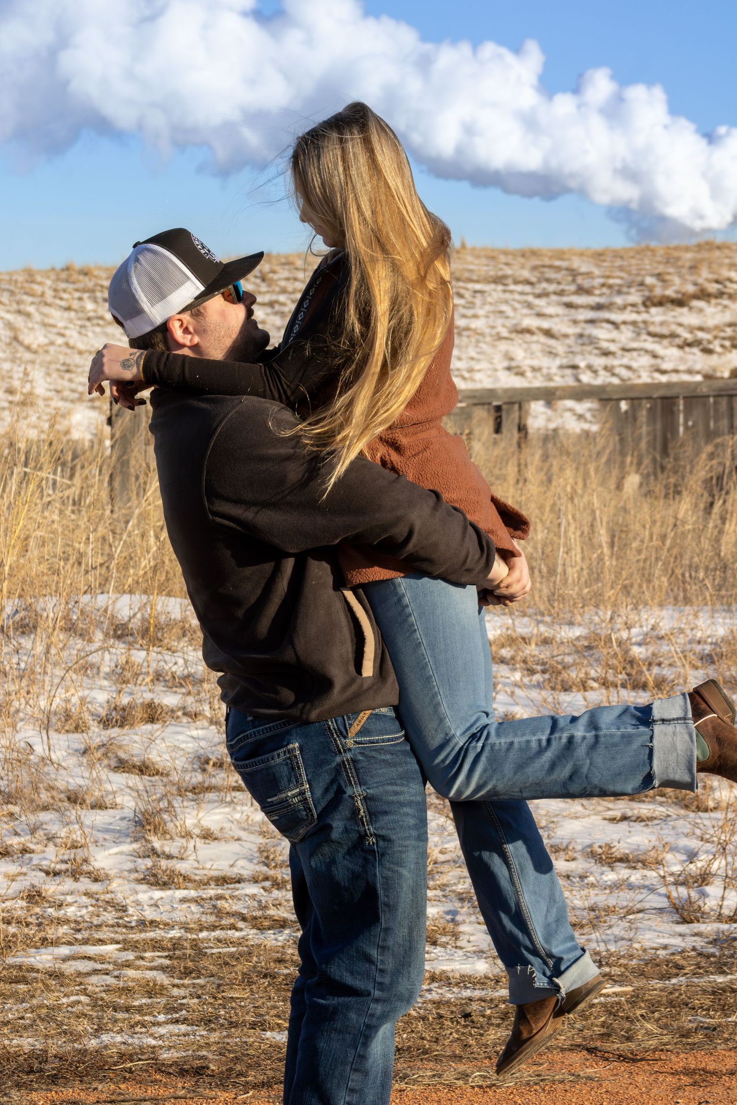 A couple embraces in a playful lift pose against a snowy winter landscape with dry grass and cloudy skies.
