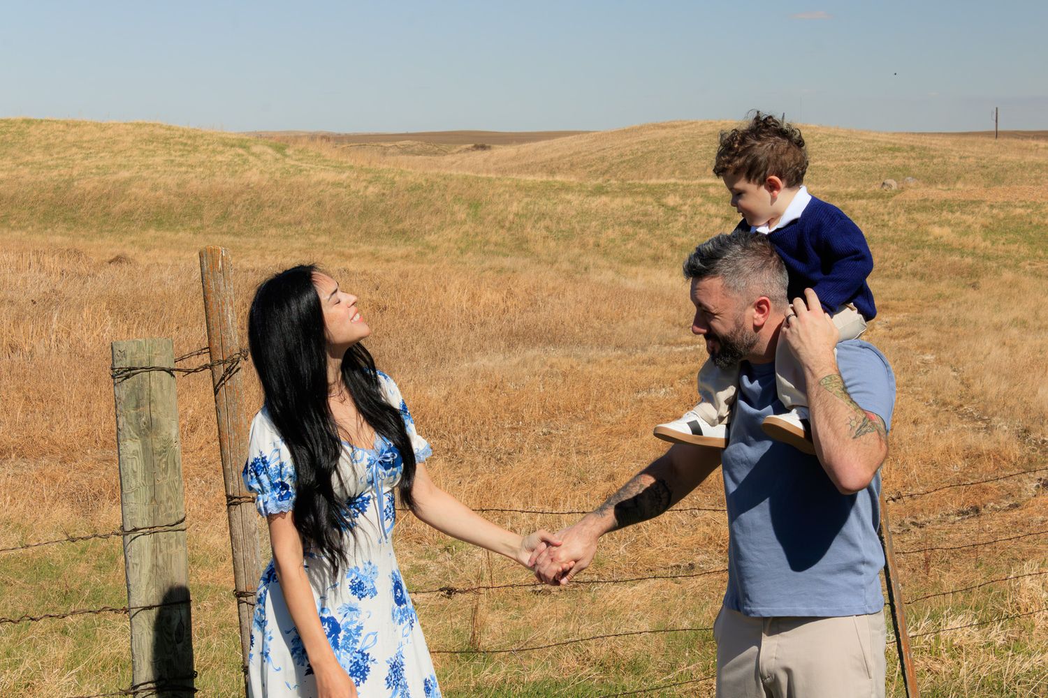 Family of three posing by a wooden fence in a golden prairie field, parents holding hands with child on dad's shoulders.