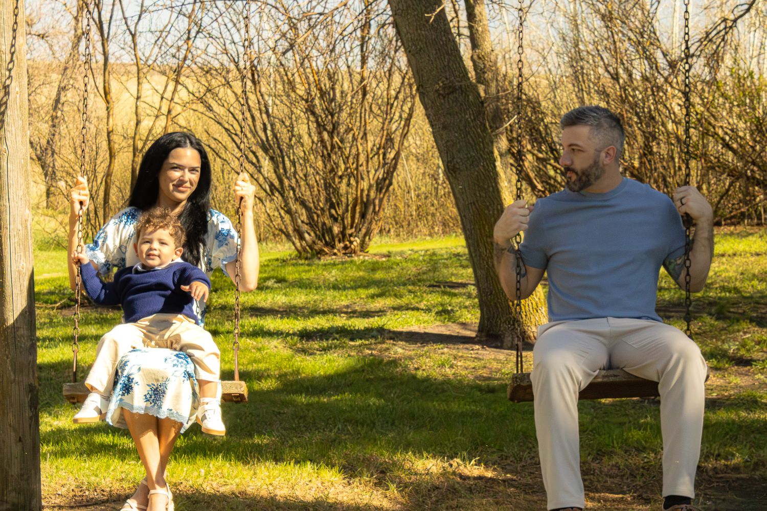 A happy family enjoys a sunny day outdoors, with a woman and child sharing a swing while a man relaxes on a nearby swing.