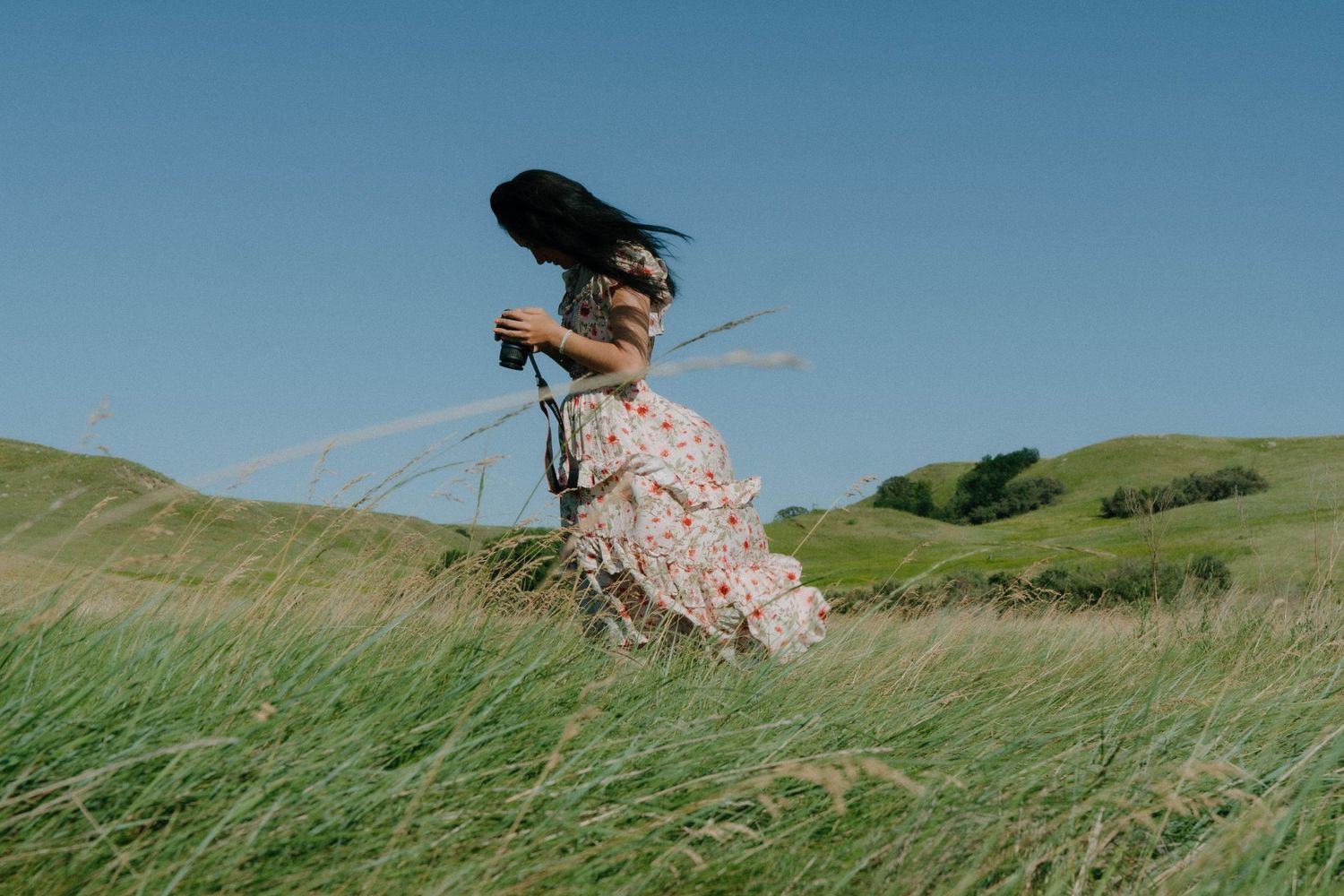 Figure in a floral dress running through tall grass on rolling hills under blue sky.