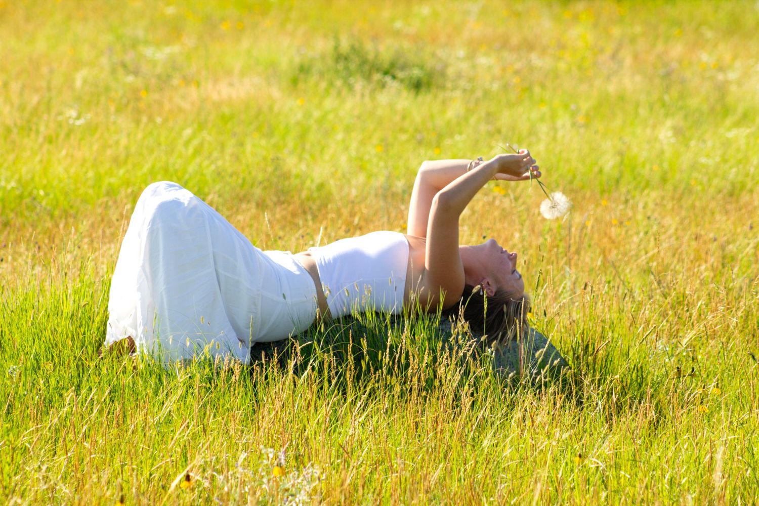Woman in white dress lying in sunny green grass field, holding a dandelion flower up toward the sky.