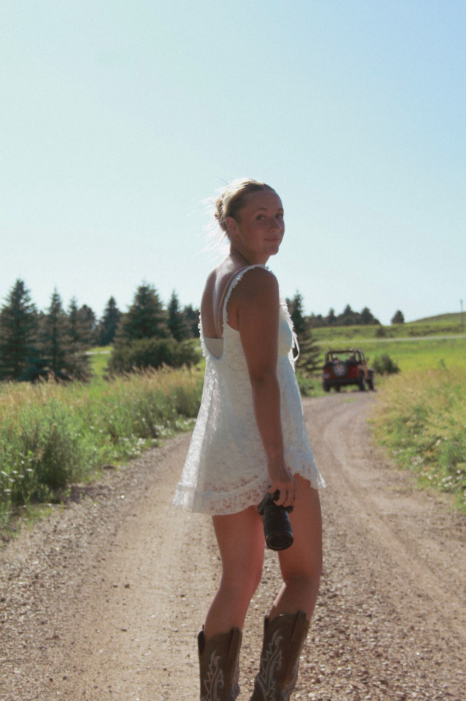 A photographer in cowboy boots and white dress stands on a rural dirt road with evergreen trees.