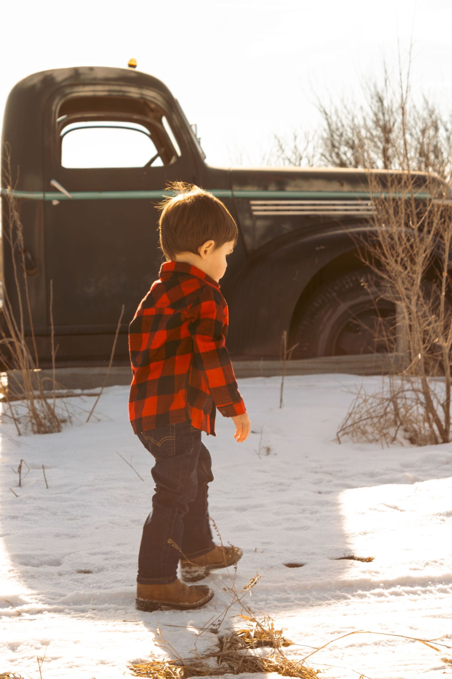 A small child in a red plaid jacket and dark pants walks through snowy terrain with vintage trucks in the background.