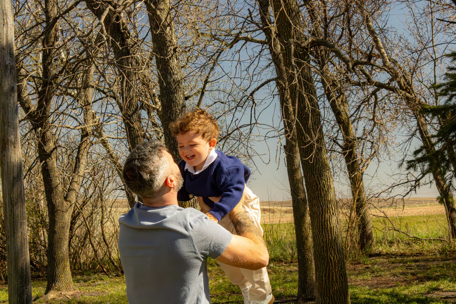 A father lifts his young son in the air outdoors among bare trees on a sunny day.