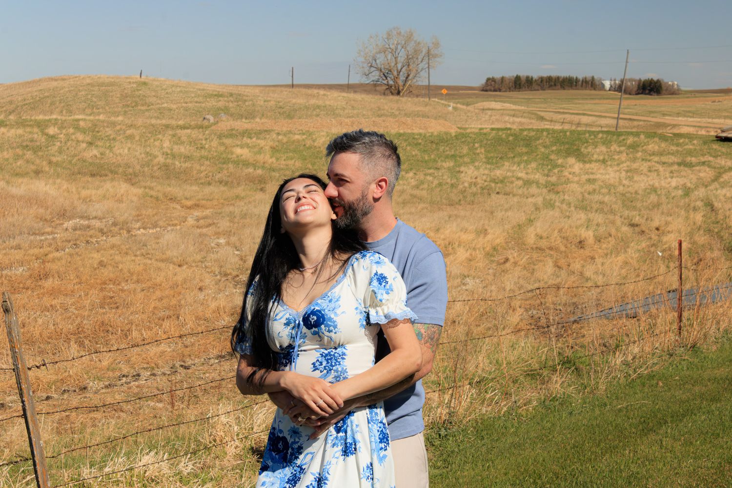 Couple embracing in a sunny rural field, woman in blue floral dress smiling as man kisses her cheek.